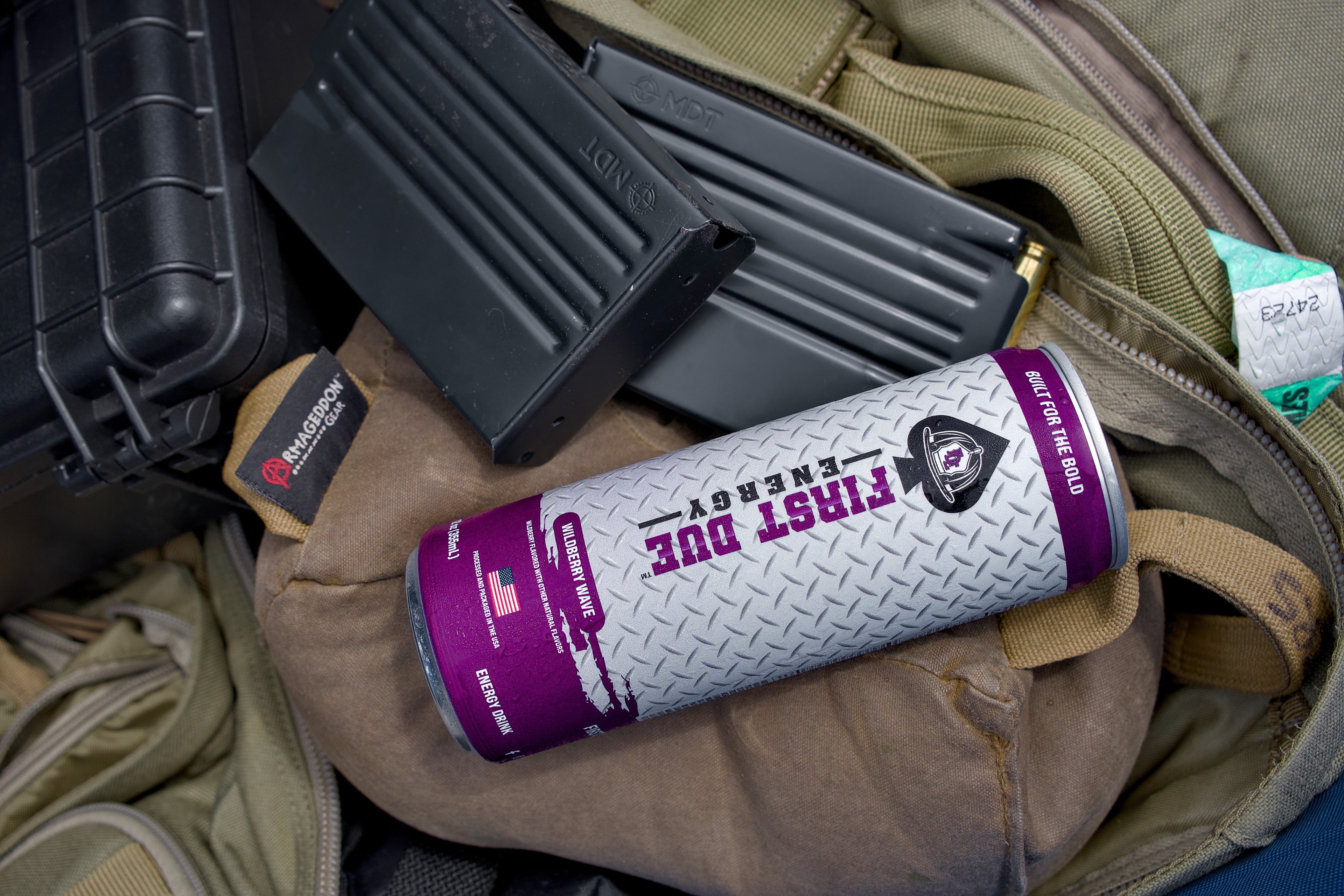 A can of First Due Energy drink lies on top of a tan backpack, surrounded by black rifle magazines and a hard case.