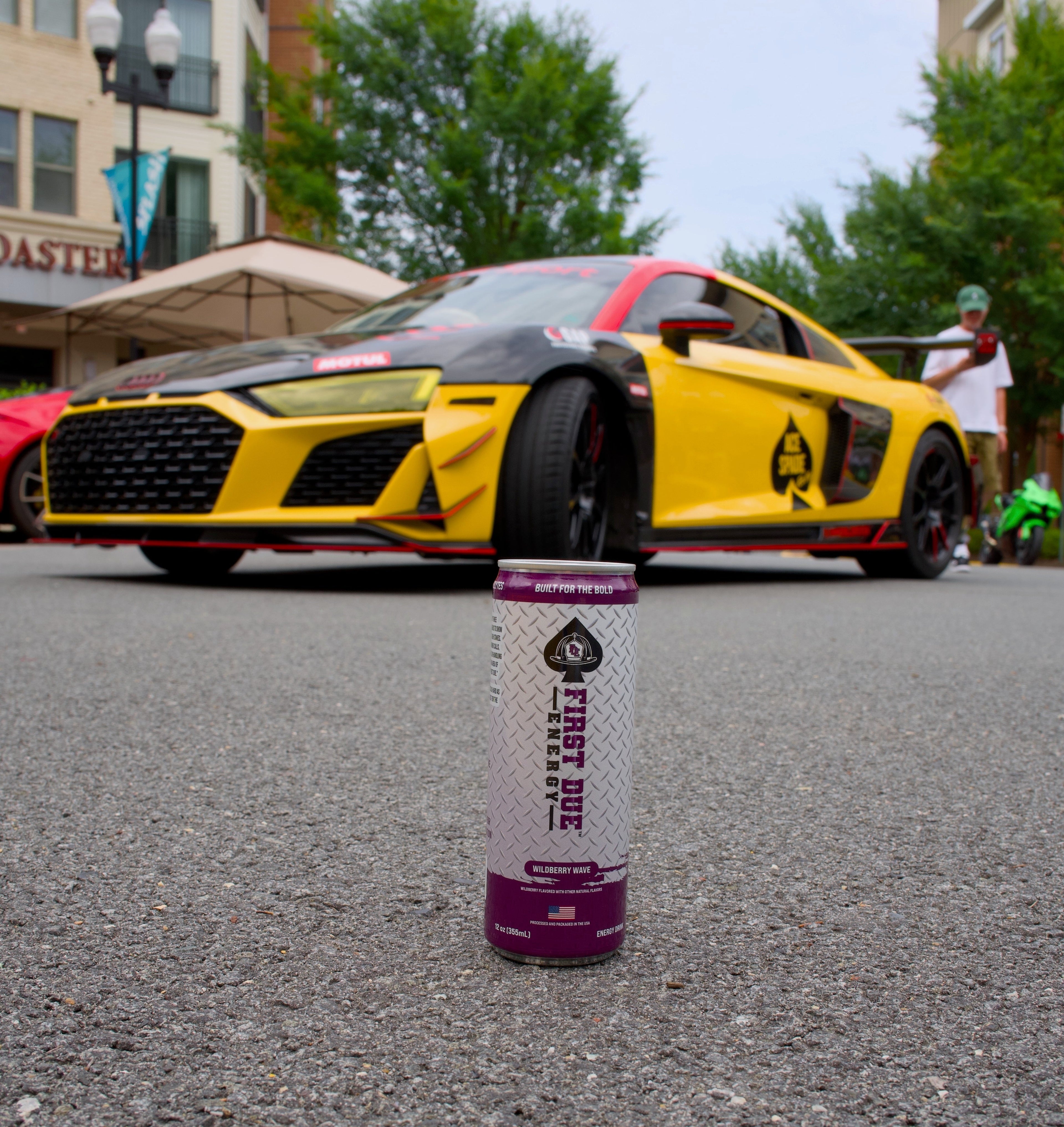 A can of First Due Energy drink is placed on the asphalt road in the foreground, with a yellow and black race car driving in the background on a city street.