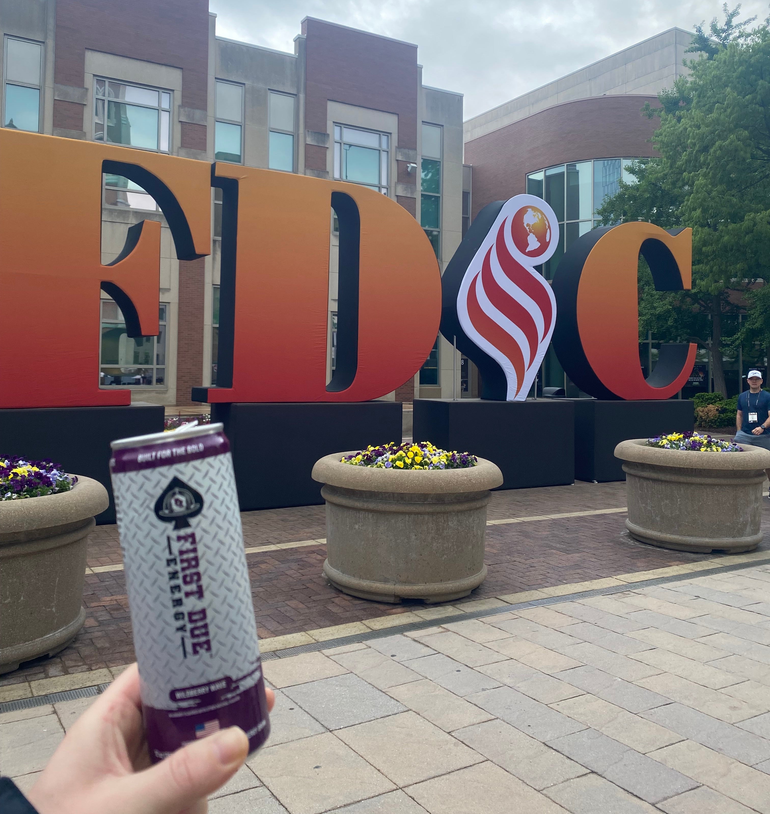 A hand holding a can of First Due Energy drink in the foreground, with a large sign spelling 'FDIC' in the background under an overcast sky, at the Fire Department Instructors Conference in Indianapolis.
