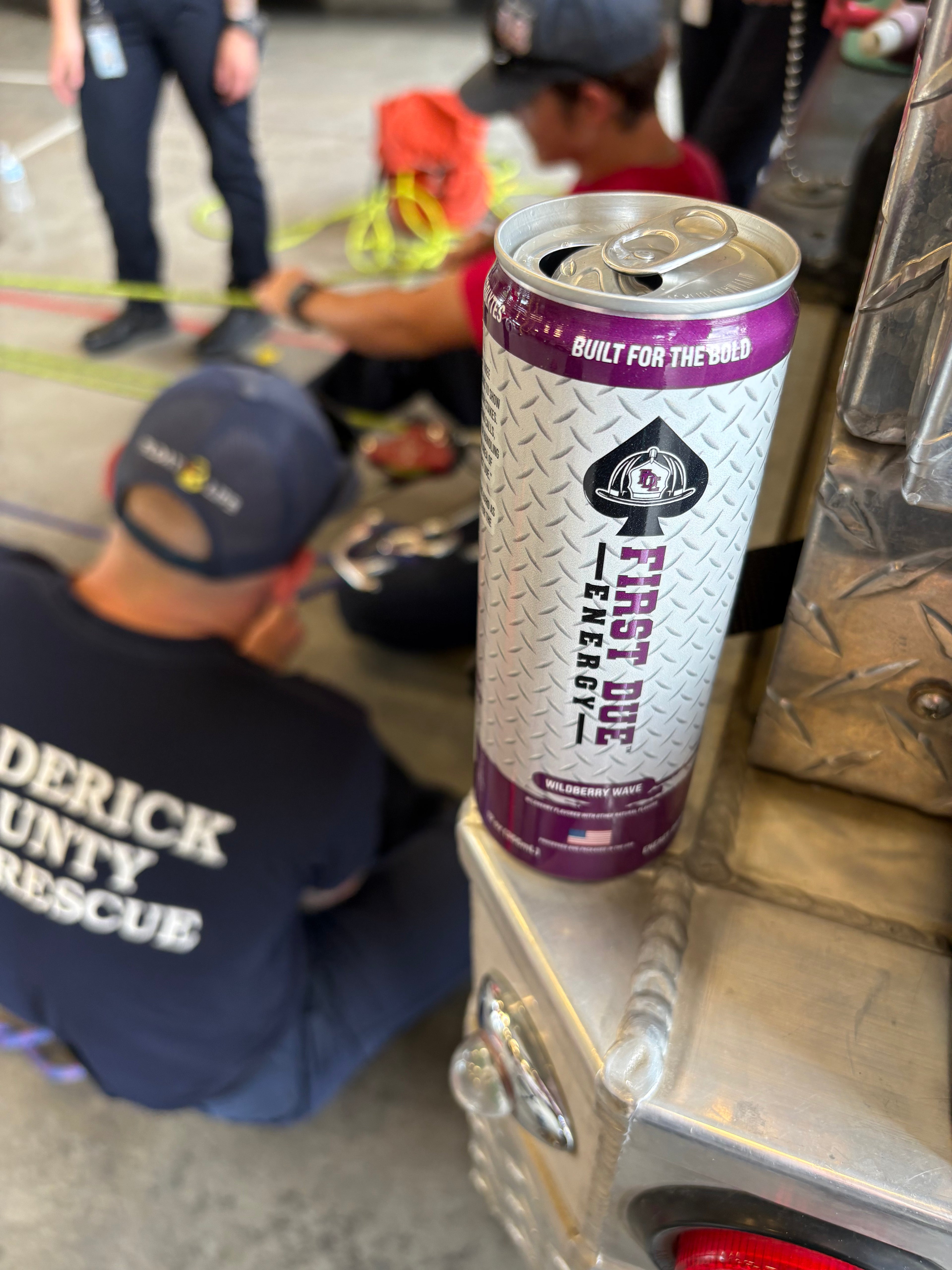 A silver and purple can of First Due Energy drink is shown on the bumper of a Frederick County Fire and Rescue Department fire truck, with a training exercise on rope techniques occurring in the background.