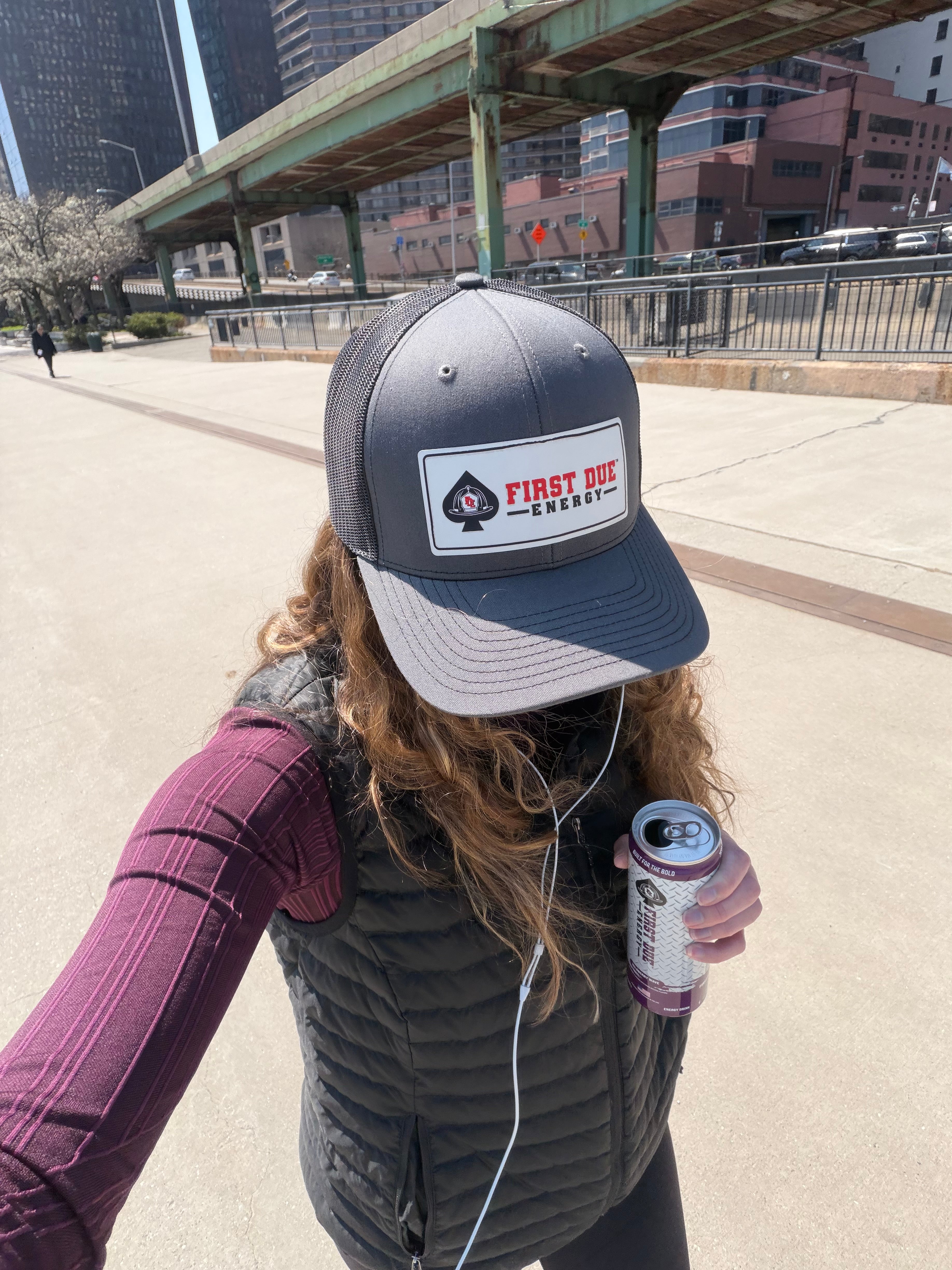 A young woman outdoors in New York City wearing a dark gray trucker-style baseball cap with a white patch that reads 'FIRST DUE ENERGY' and holds a First Due Energy beverage can. The background shows an urban walkway with buildings and a highway overpass.