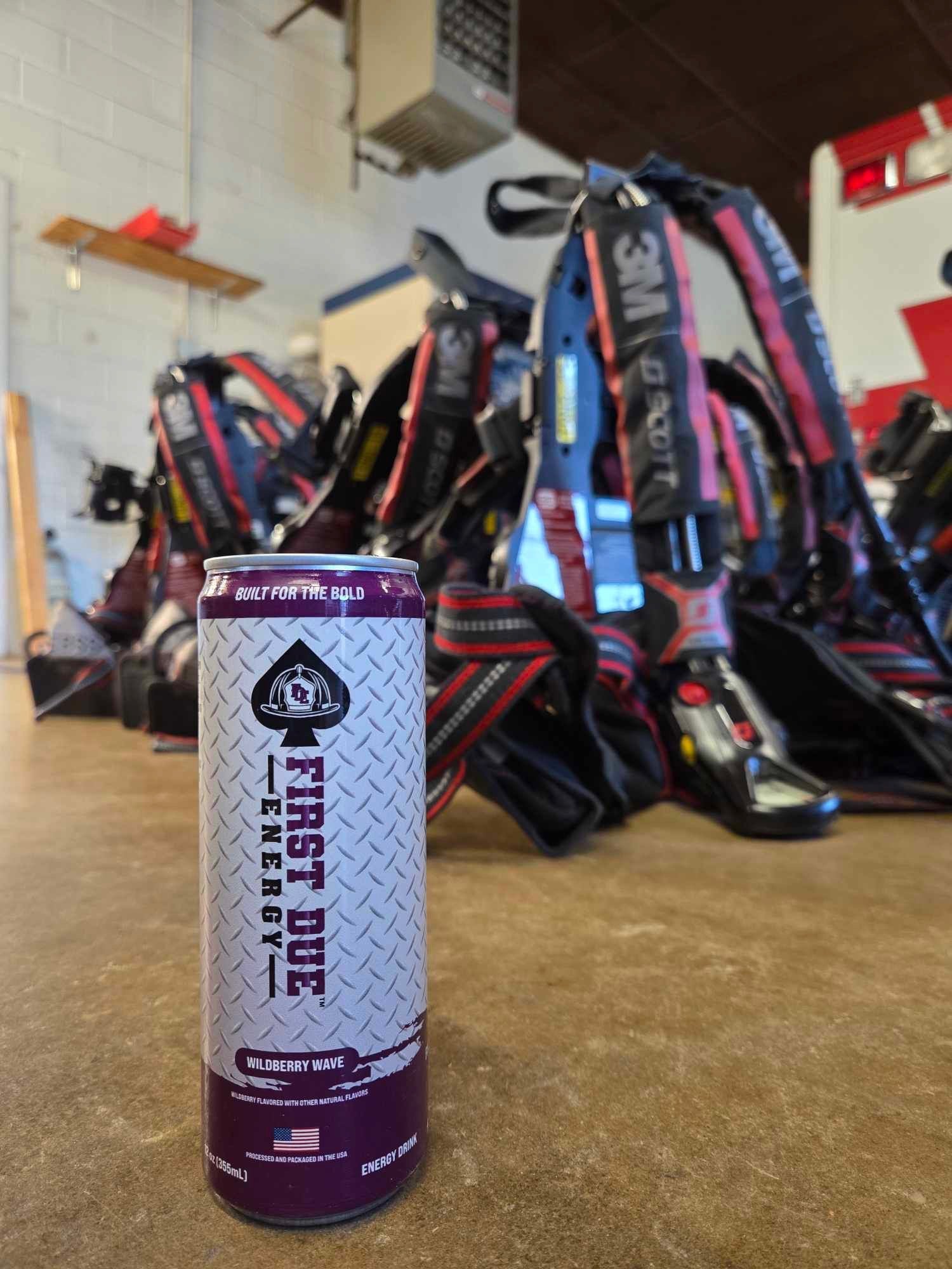 A can of First Due Energy drink placed in the foreground on a concrete floor with firefighting SCBA air packs blurred in the background.
