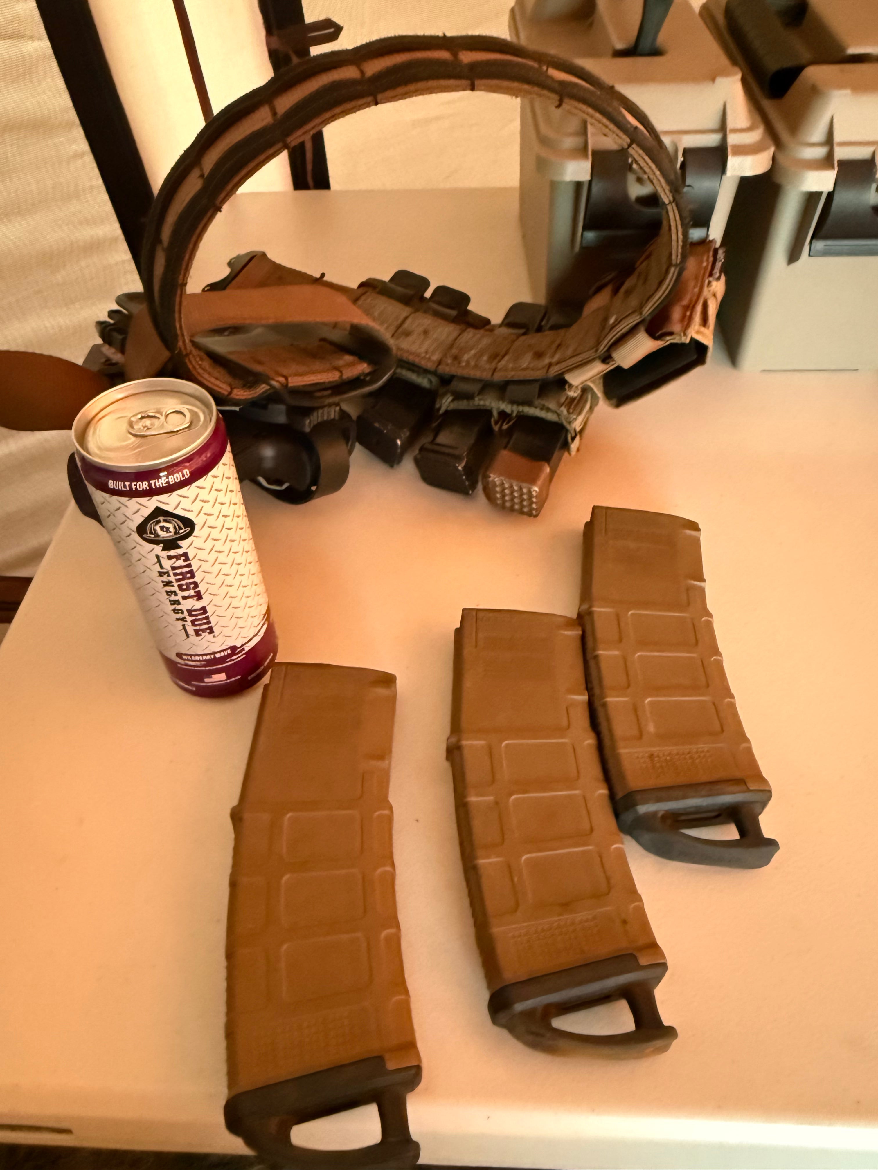 Three tan polymer magazine-like accessories with black end pieces arranged on a white table, alongside a can of energy drink and a brown leather-like curved holster in the background.