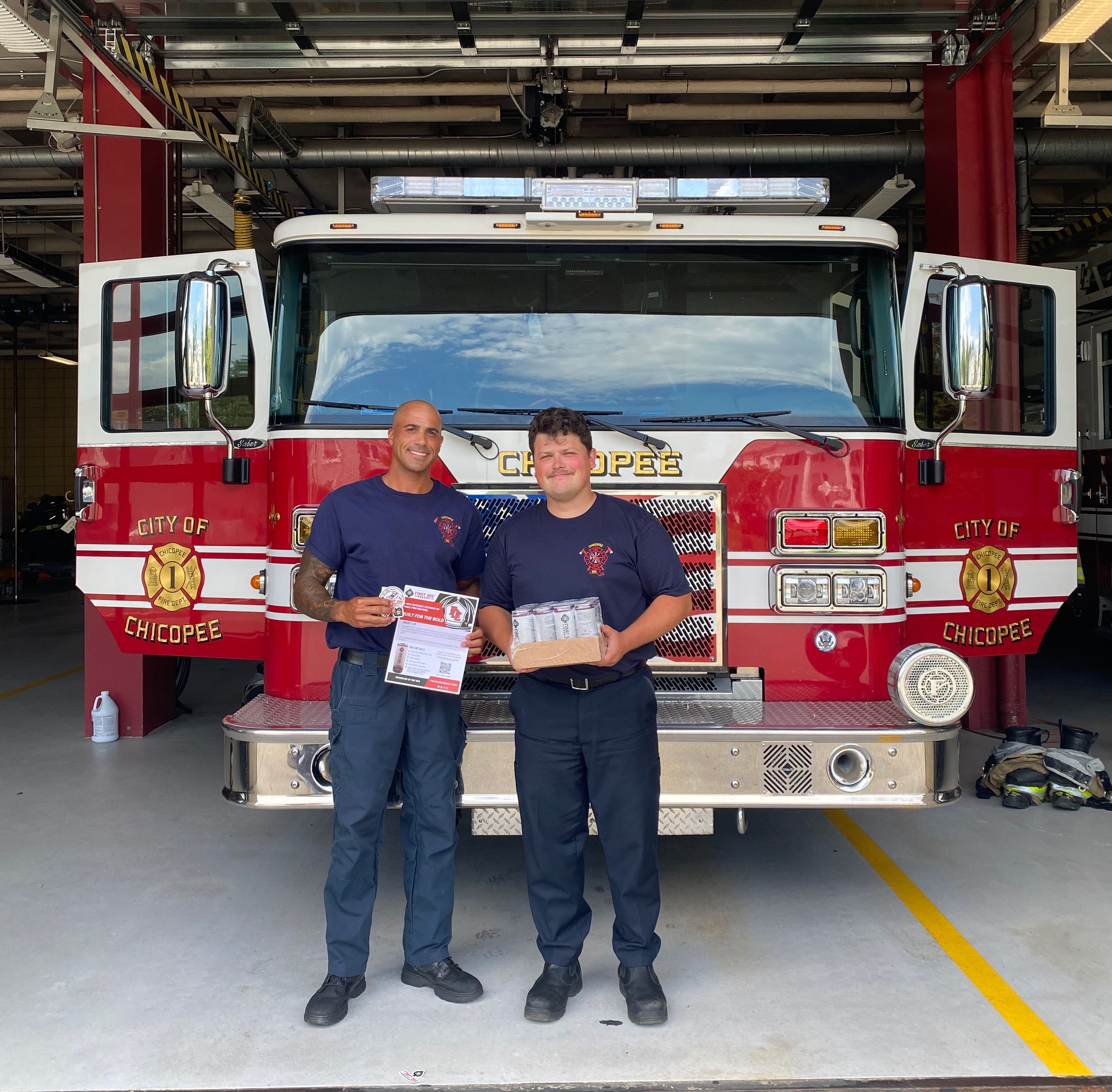 Two firefighters standing in front of a red and white City of Chicopee, MA, fire truck inside a fire station garage, holding a First Due Energy flyer and case of drinks.