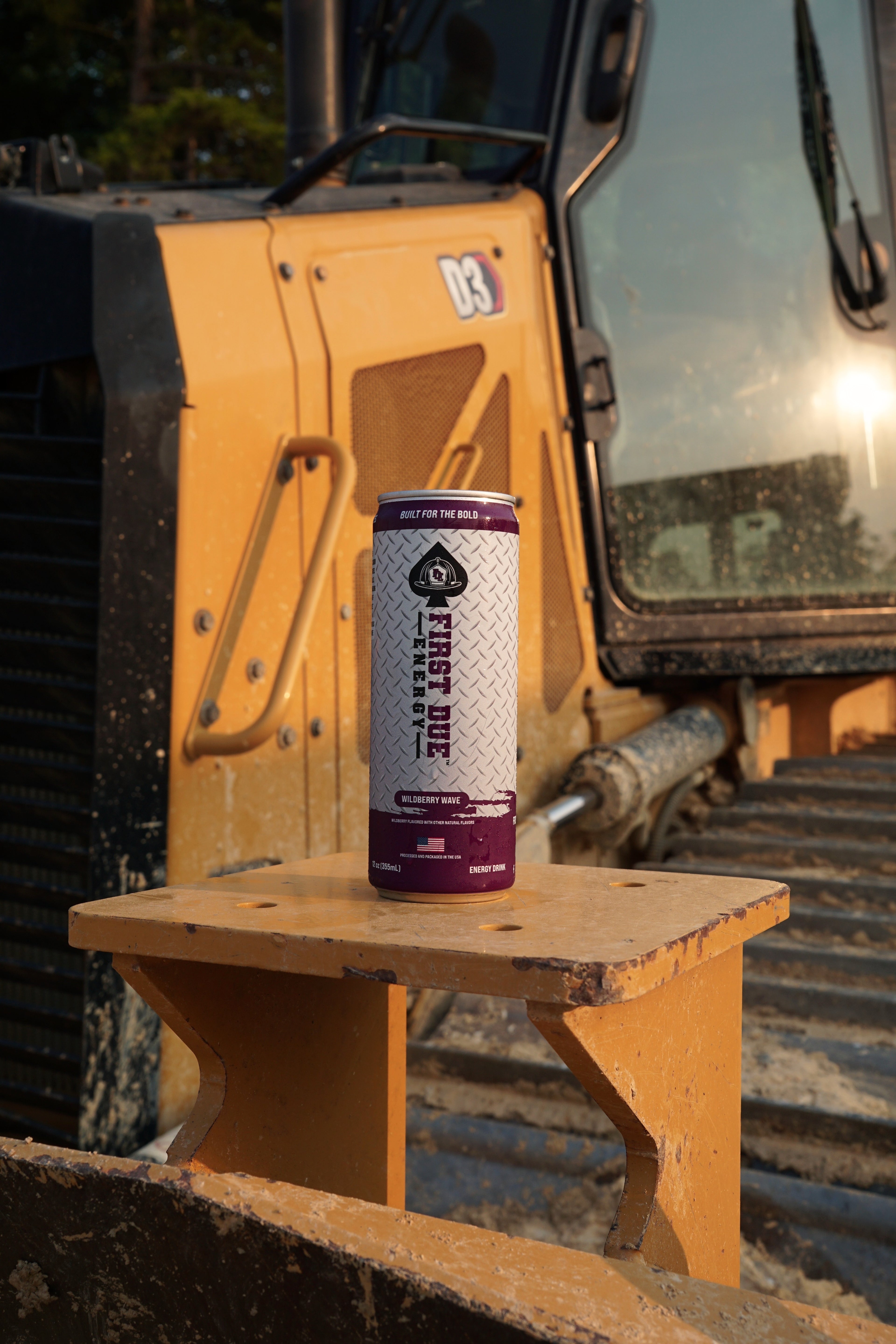A single can of First Due Energy sits on a the edge of a yellow construction vehicle, with the product label clearly visible against a dusty outdoor backdrop.