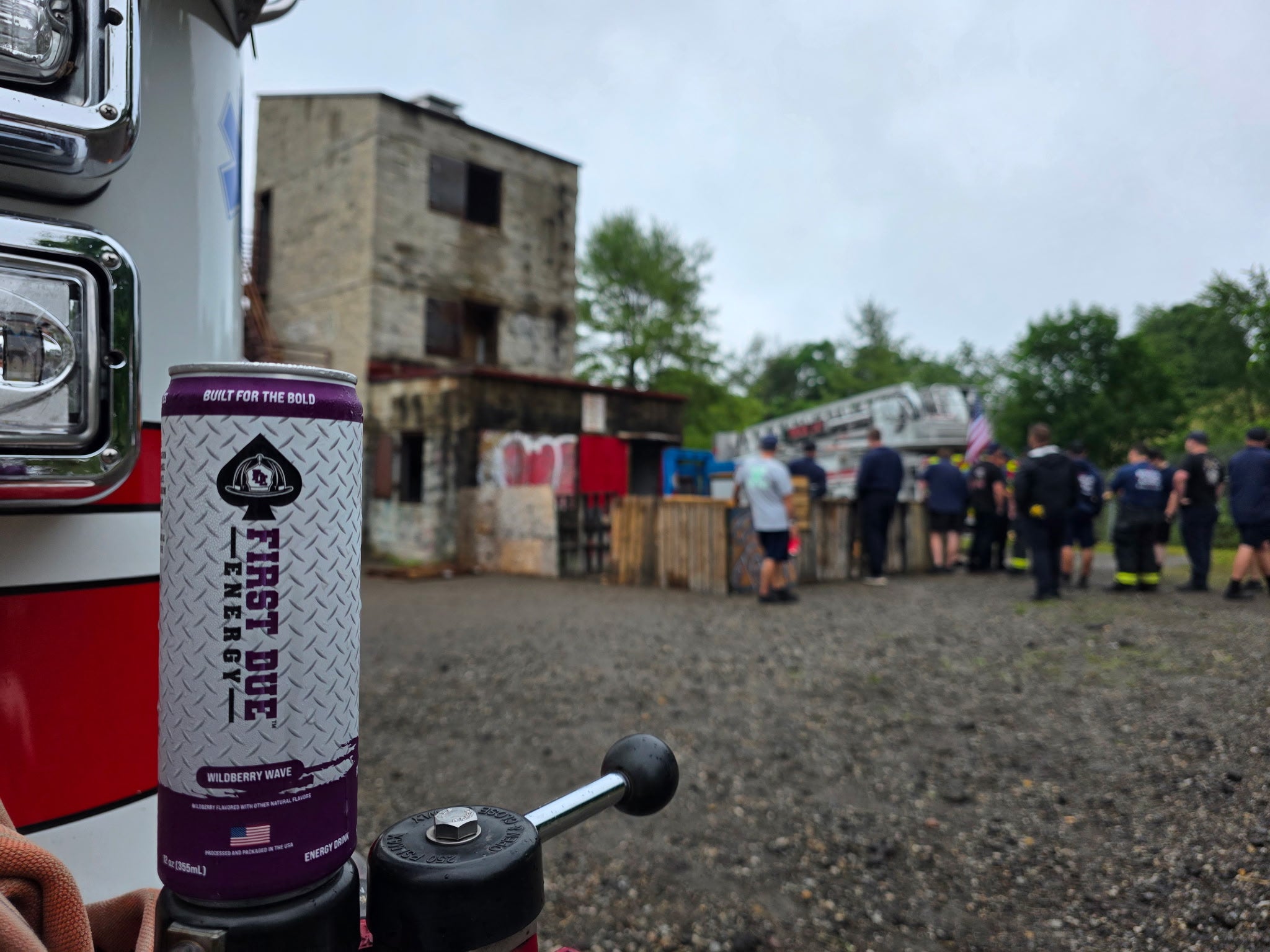 A close-up of a First Due Emergy beverage can in the foreground, with a fire truck bumper visible on the left and a group of firefighters standing near a ladder truck in the blurred background doing firefighting operations training.
