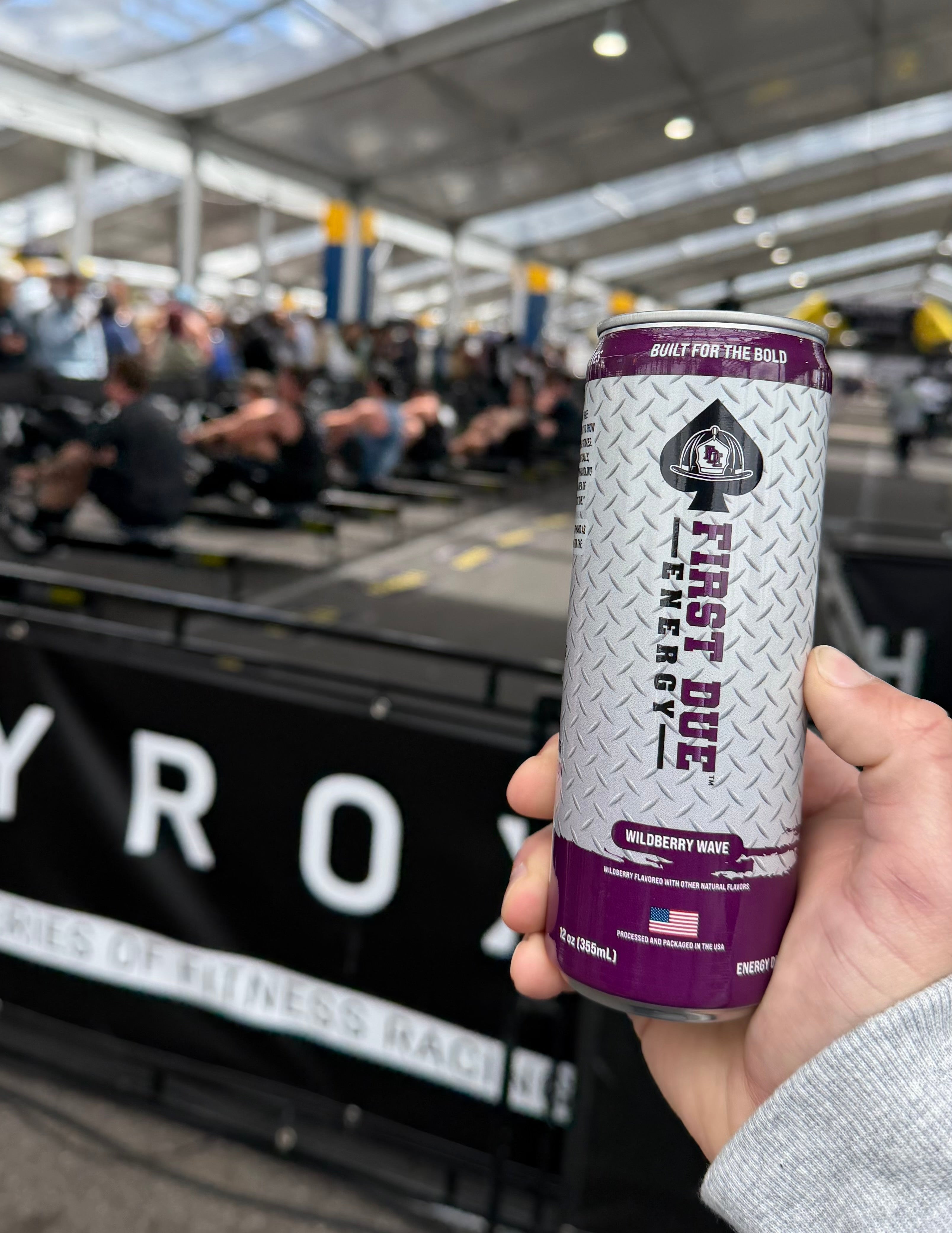 A hand holds a can of First Due Energy drink at a Hyrax competition, with people working out on rowing machines in the background.
