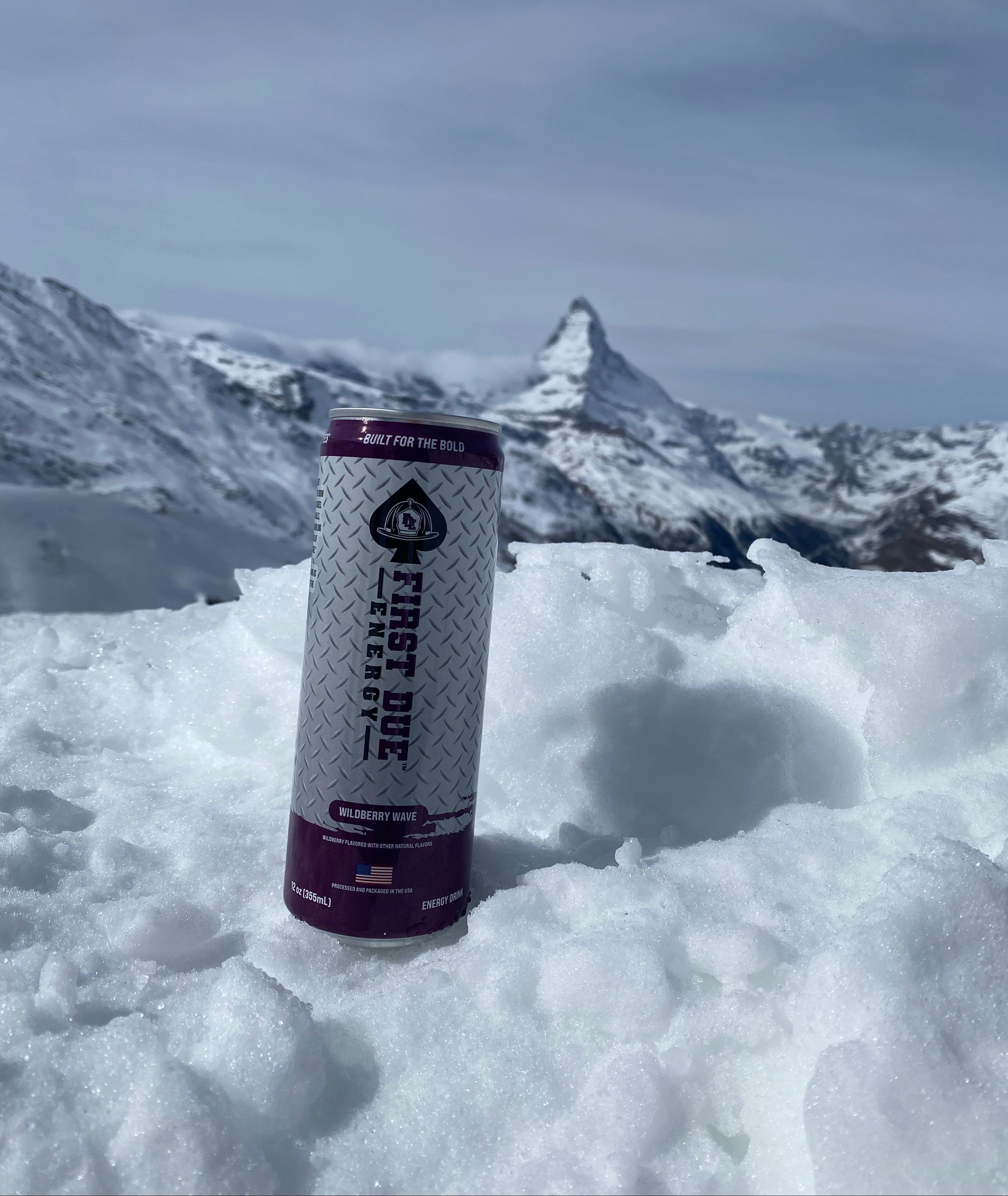 A can of First Due Energy drink placed in the snow on the slopes at Zermatt in Switzerland, with the Matterhorn and blue sky in the background.