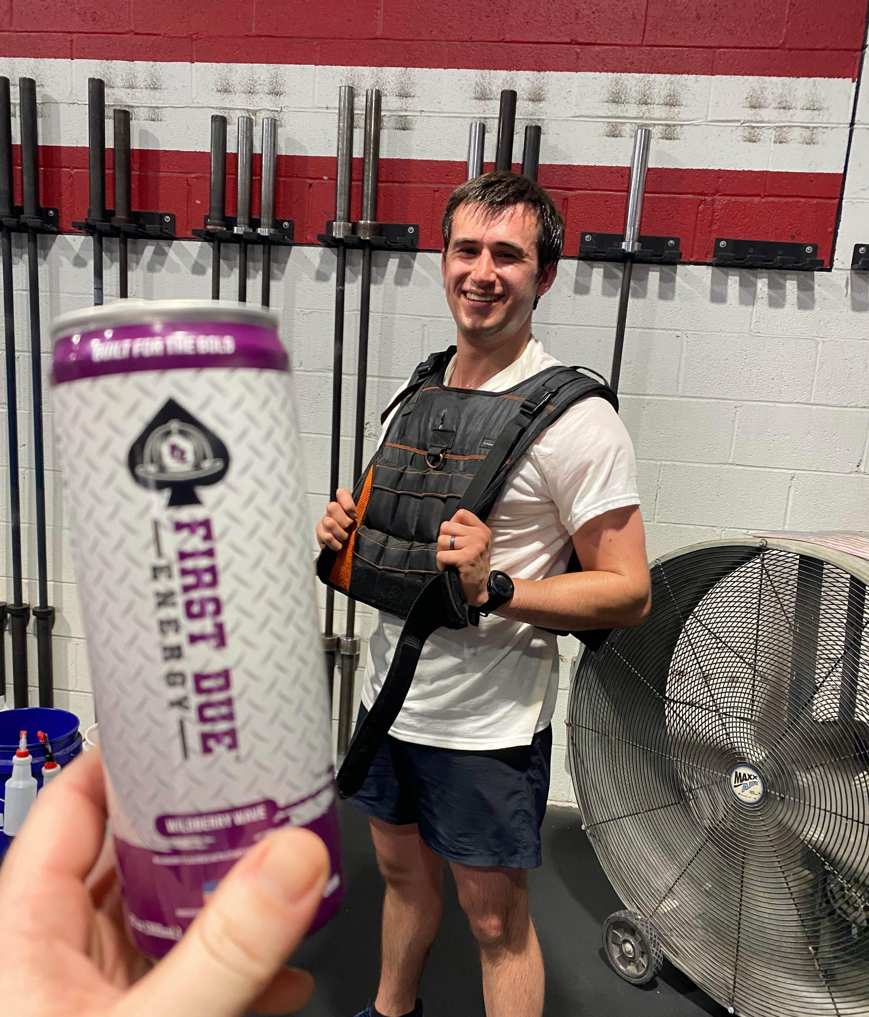 A person in a gym wearing a weight vest in front of a wall of barbells, with another person's hand holding a First Due Energy can in the foreground.