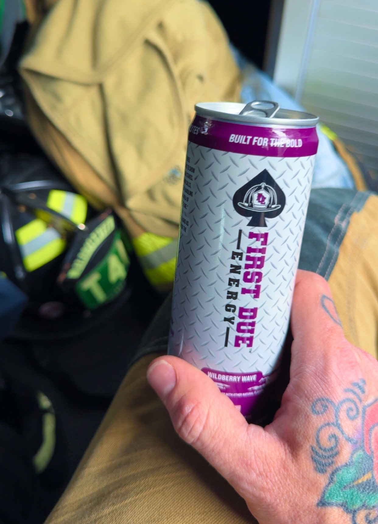 A hand with a tattoo holds a can of First Due Energy drink in the back of a fire engine, with bunker gear and a helmet in the background.